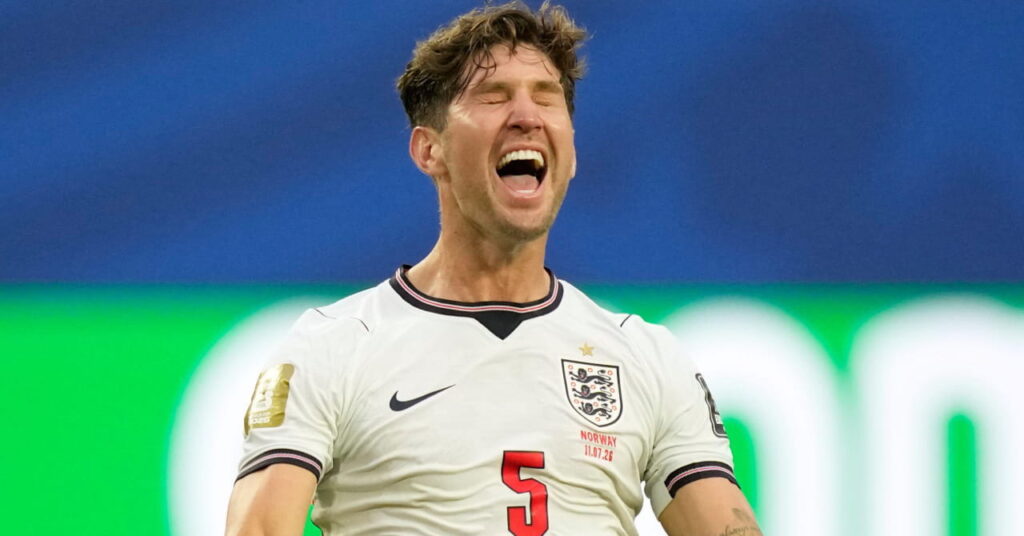 Fotballspiller John Stones kneler på en bane med armene utstrakt smilende og ropte i feiring mens han spilte i Englands landslagsdrakt i hvitt med blå og røde detaljer under VM 2026