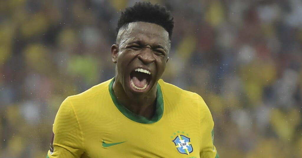 Et bilde av fotballspiller Vinicius Junior som roper i feiring og smiler mens han har på seg en skjorte i Brasils fotballag på et stadion. Skjorten er gul med grønn kant og har en grønn Nike-flått og Brasil-merke på brystet