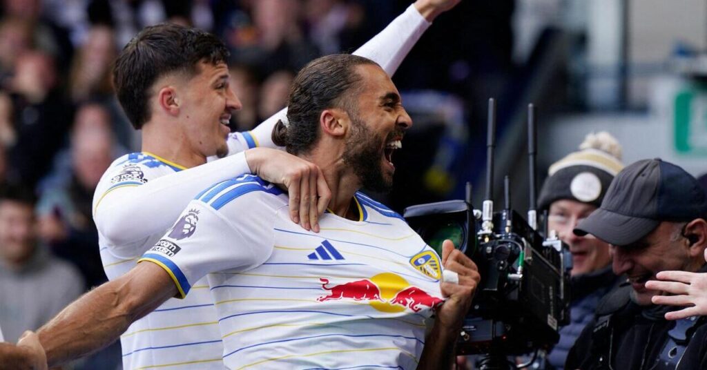 Fotballspiller Dominic Calvert-Lewin ser til venstre på et stadion og smiler og roper for å feire et mål mens han spilte for Premier League-laget Leeds United i en hvit drakt med blå kant og gule striper. Calvert Lewin er i nærheten av to kameraoperatører og blir omfavnet av en lagkamerat bak seg med Red Bull-logoen i rødt og gult, Adidas-logoen i blått og klubbmerket på brystet.